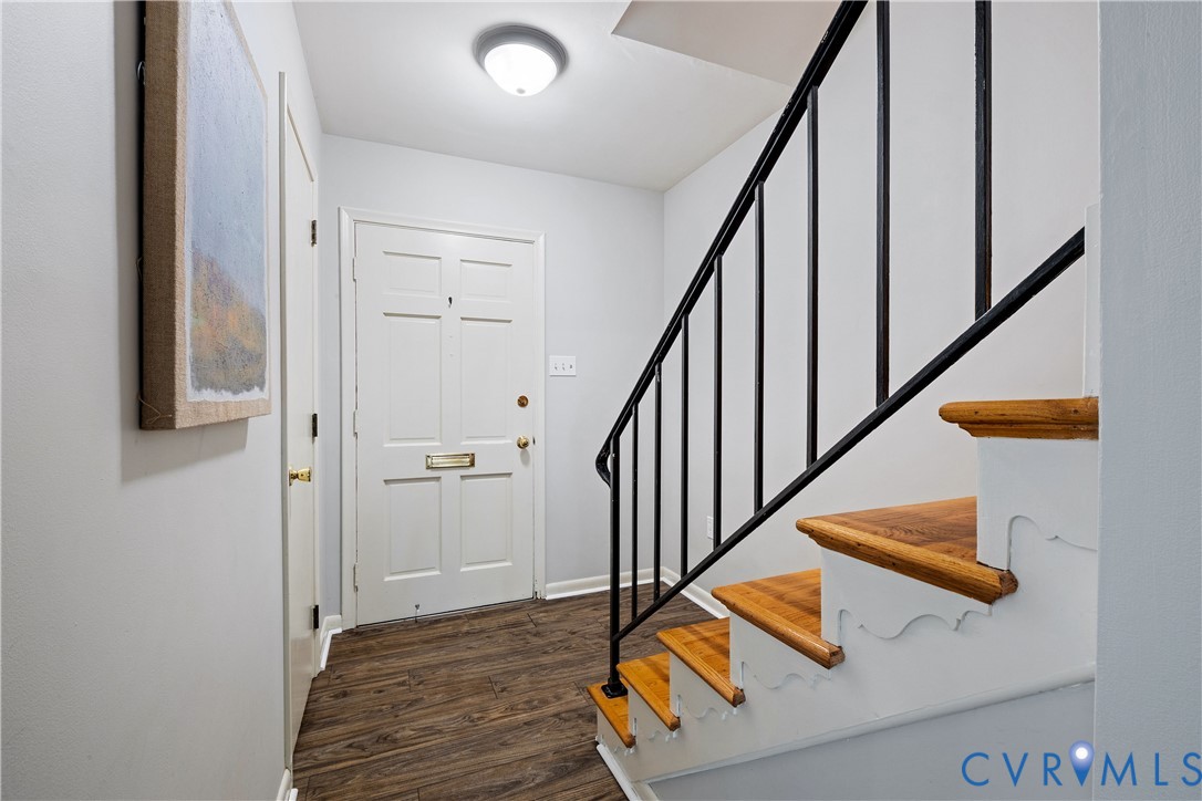 Foyer featuring wood-finish flooring, a white panel door with brass hardware, a staircase with wood treads and decorative risers, a black metal handrail, and a flush-mount ceiling light fixture Foyer featuring wood-finish flooring, a white panel door with brass hardware, a staircase with wood treads and decorative risers, a black metal handrail, and a flush-mount ceiling light fixture