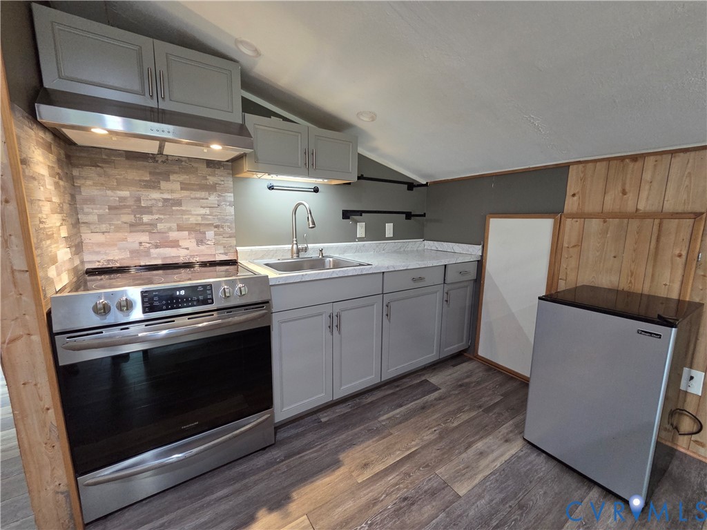Kitchen featuring wood-finish flooring, grey cabinetry, stainless steel range with overhead vent hood, stone-style tile backsplash, and light-toned countertop with integrated sink