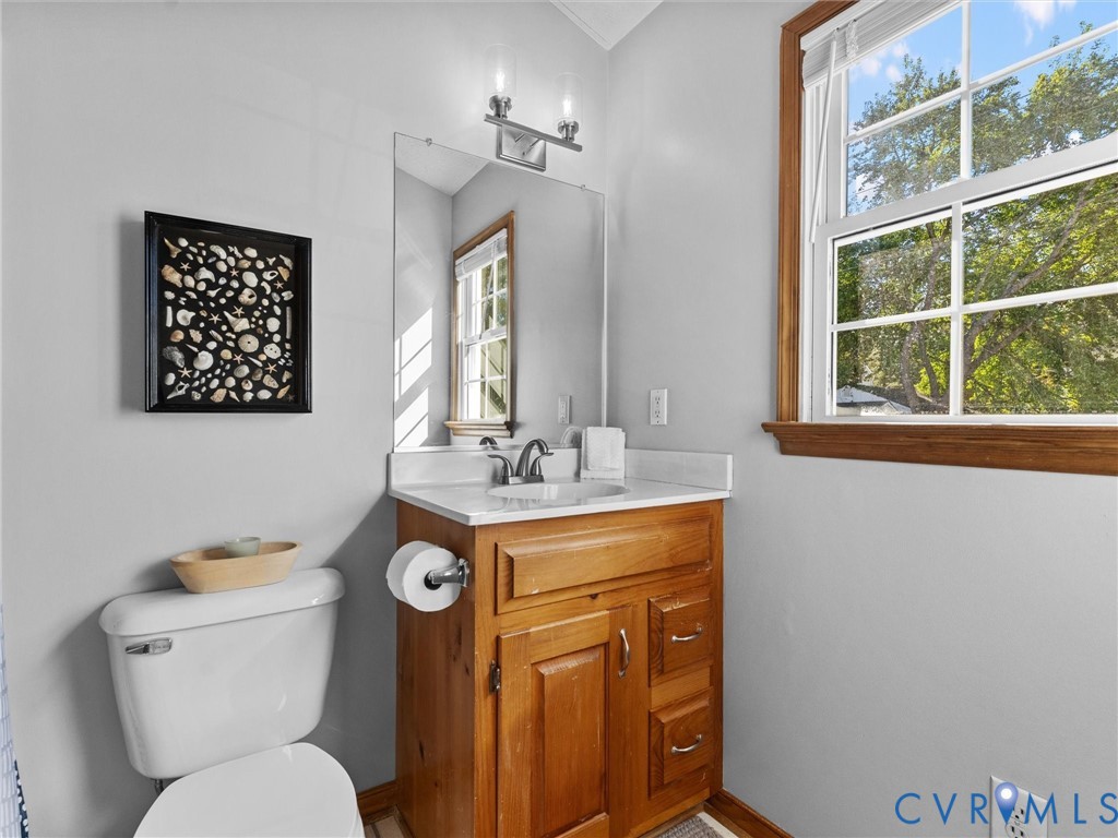 Bathroom featuring a natural wood-finish vanity, light-toned countertop with an integrated sink, polished metal dual-handle faucet, wall-mounted vanity light, and a double-hung window with wood trim