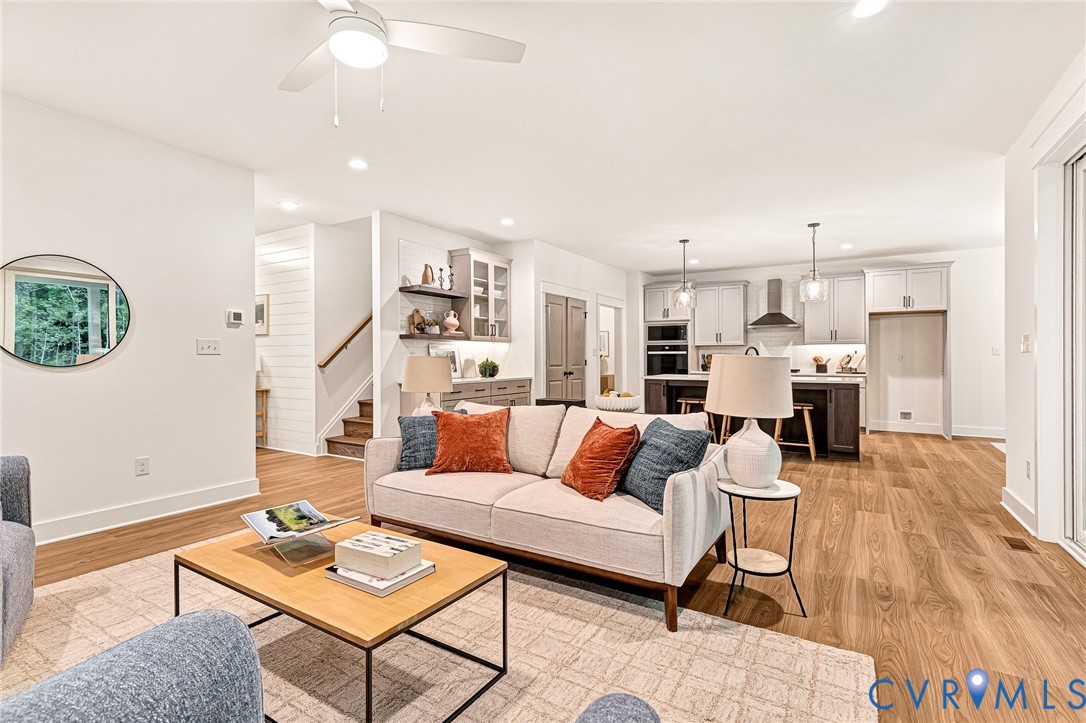 Open concept living area featuring wood-finish flooring, recessed lighting, a modern ceiling fan, built-in cabinetry with open shelving, and a kitchen island with pendant lighting