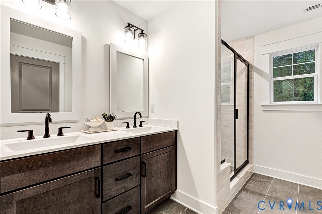 Dual-sink vanity with dark wood-finish cabinetry, light-toned countertop, matte black hardware, and framed mirrors with overhead industrial-style lighting, complemented by a walk-in shower with black-frame glass enclosure and large-format tile flooring