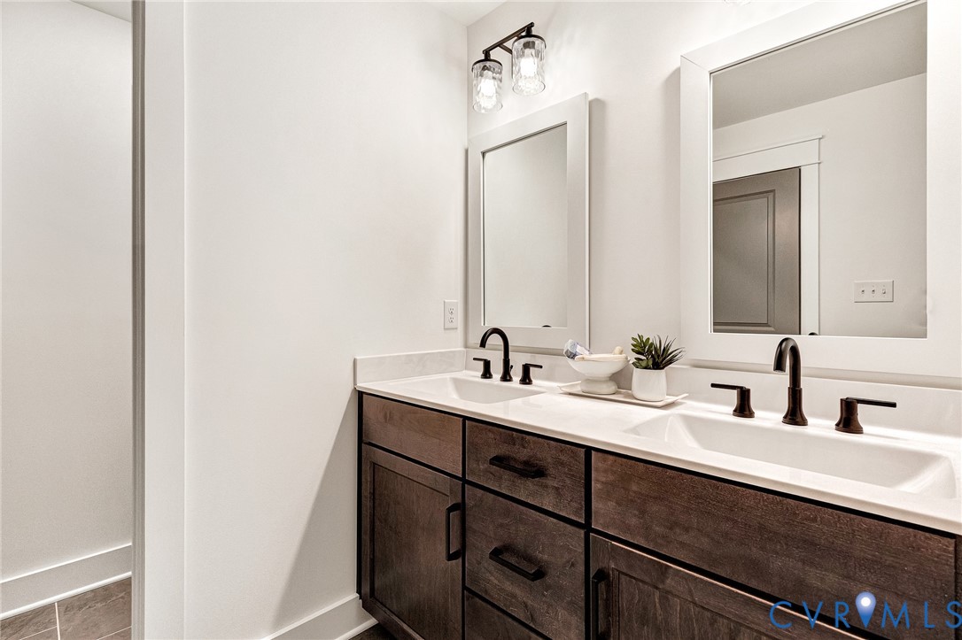 Dual-sink vanity featuring dark wood-finish cabinetry, a white countertop, rectangular under-mount sinks, and matte black hardware with matching wall-mounted lighting