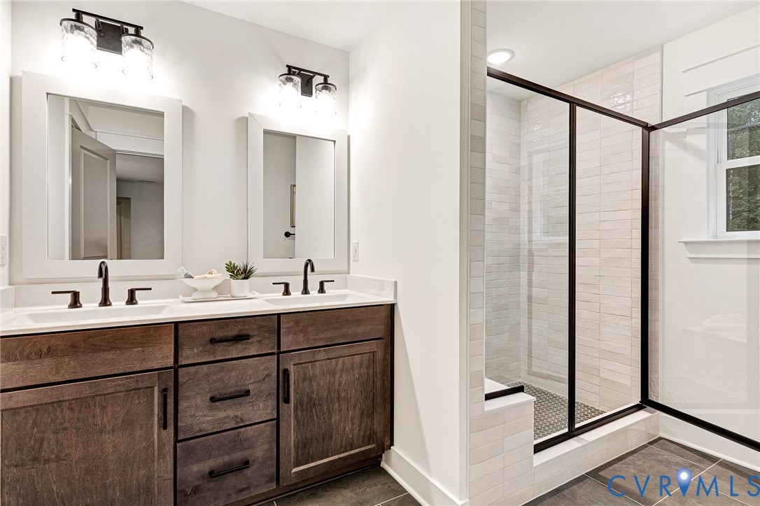 Double vanity featuring stained wood cabinetry, quartz countertops, matte black fixtures, and a walk-in shower with a glass enclosure and tiled surround