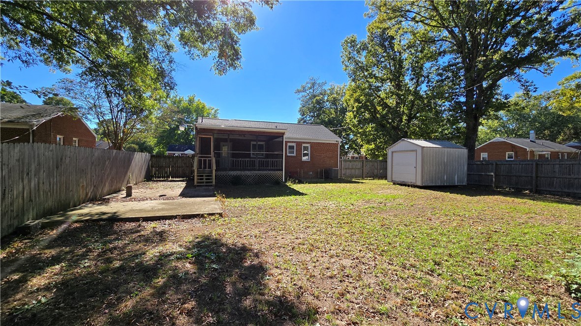 Rear exterior featuring a brick facade, covered porch with wooden railings, concrete patio, detached metal storage shed, and full wood privacy fencing