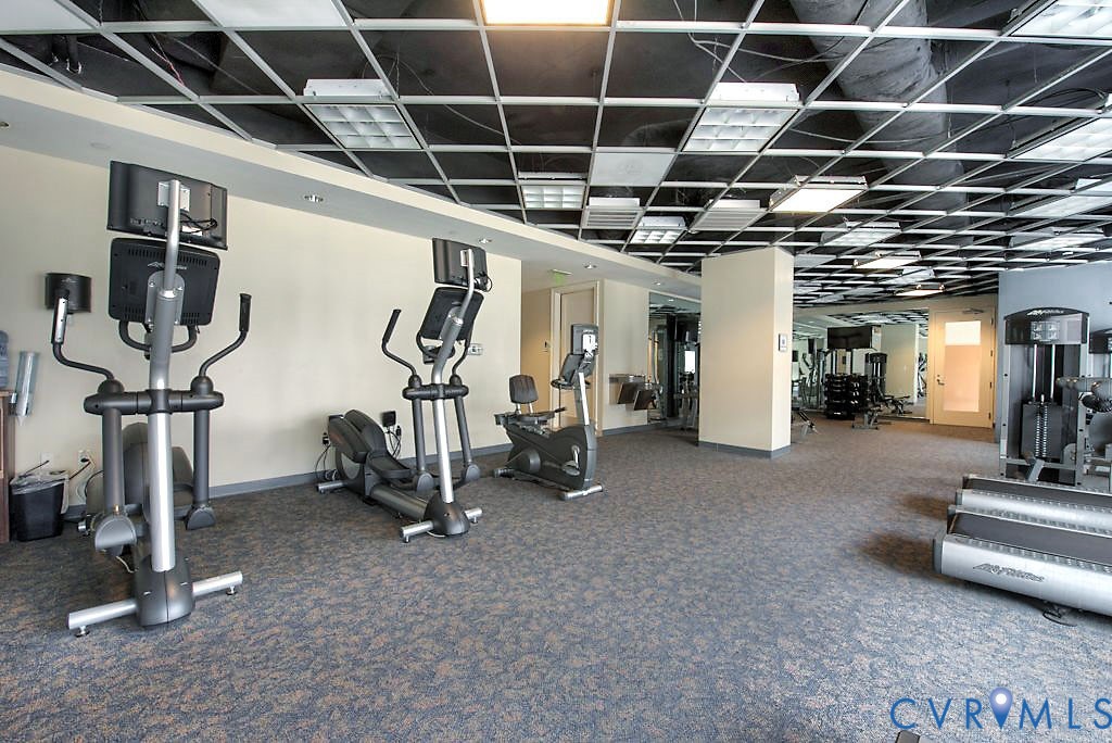 Fitness space featuring a suspended grid ceiling with integrated lighting, neutral wall finishes, patterned carpeting, and built-in mirrored walls