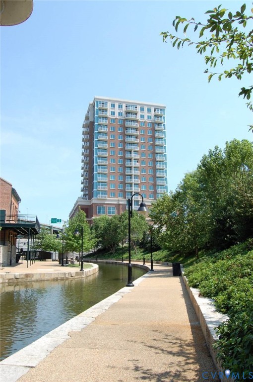 High-rise residential building featuring a brick exterior with glass-railed balconies, situated along a paved pedestrian canal path