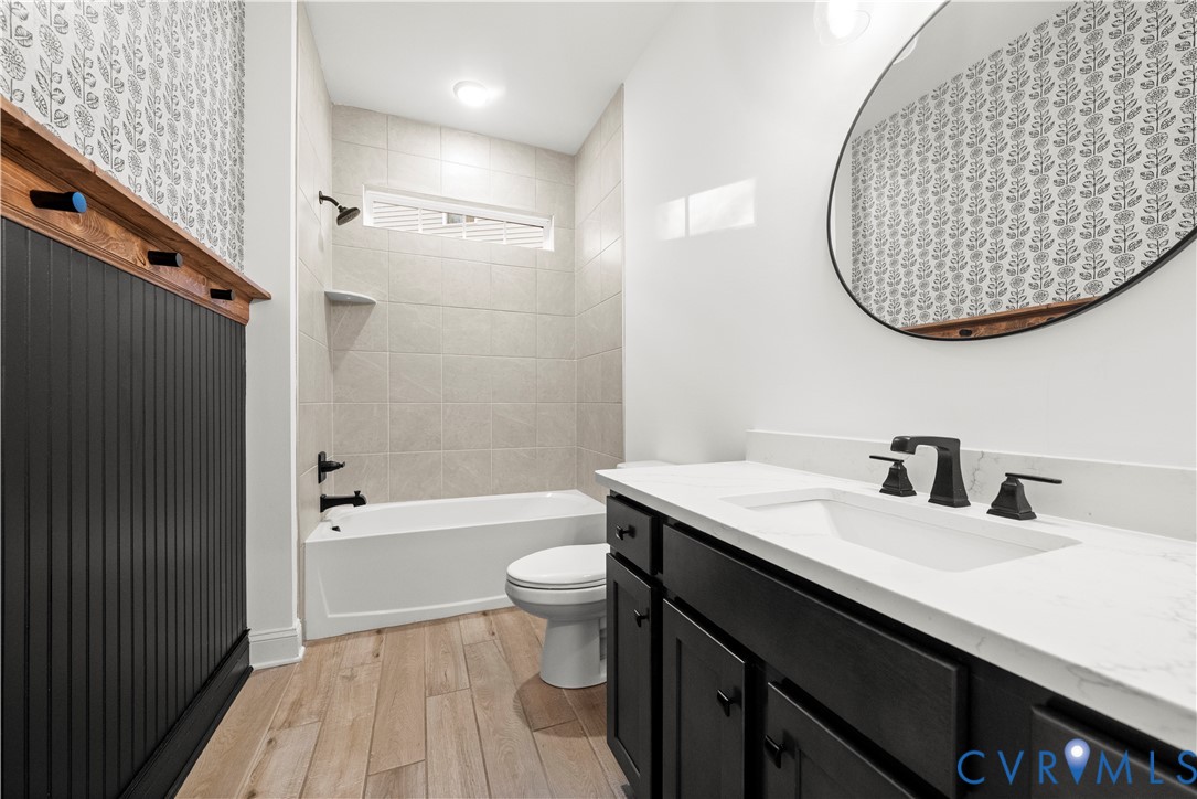 Bathroom featuring wood-finish flooring, a vanity with a light-toned countertop, matte black hardware, vertical wainscoting, and an oval wall-mounted mirror