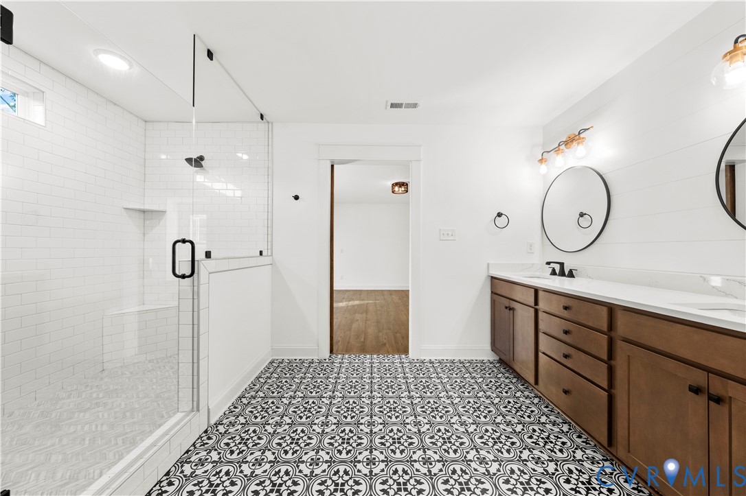 Bathroom featuring a double-sink vanity with wood cabinetry, a glass-enclosed shower with subway tile surround, patterned tile flooring, and round wall-mounted mirrors
