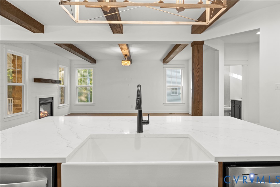 Kitchen featuring a large waterfall-edge countertop, white farmhouse sink, matte black gooseneck faucet, exposed wooden ceiling beams, and a fireplace with a wooden mantel