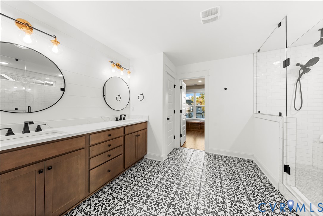 Double vanity with wood-finish cabinetry, patterned floor tile, glass-enclosed shower, round wall-mounted mirrors, and matte black hardware