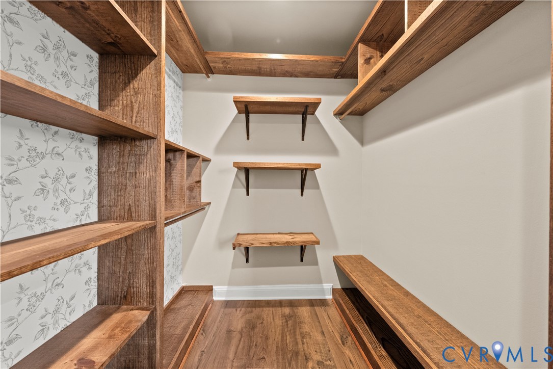 Walk-in storage space featuring built-in wood-finish shelving, wall-mounted floating wood shelves, patterned wallpaper accent, and wood-finish flooring