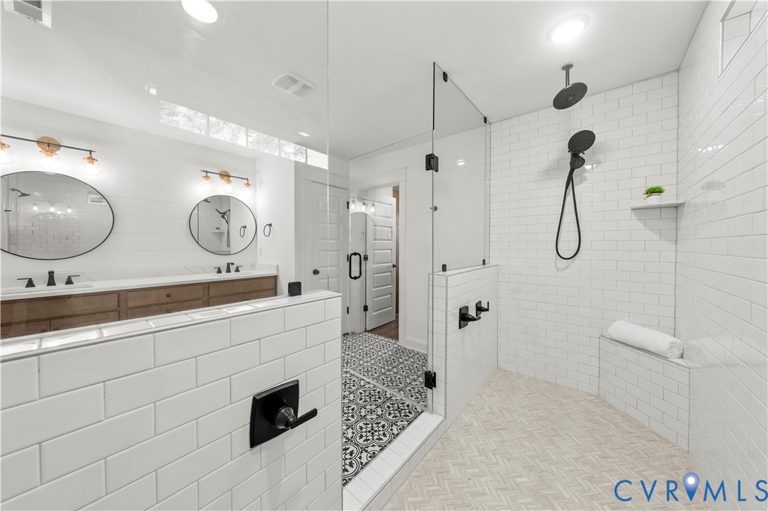 Bathroom featuring a double vanity with wood-finish cabinetry, white subway tile shower enclosure with built-in bench, matte black rainfall showerhead with handheld sprayer, glass shower partition, and decorative patterned floor tiling