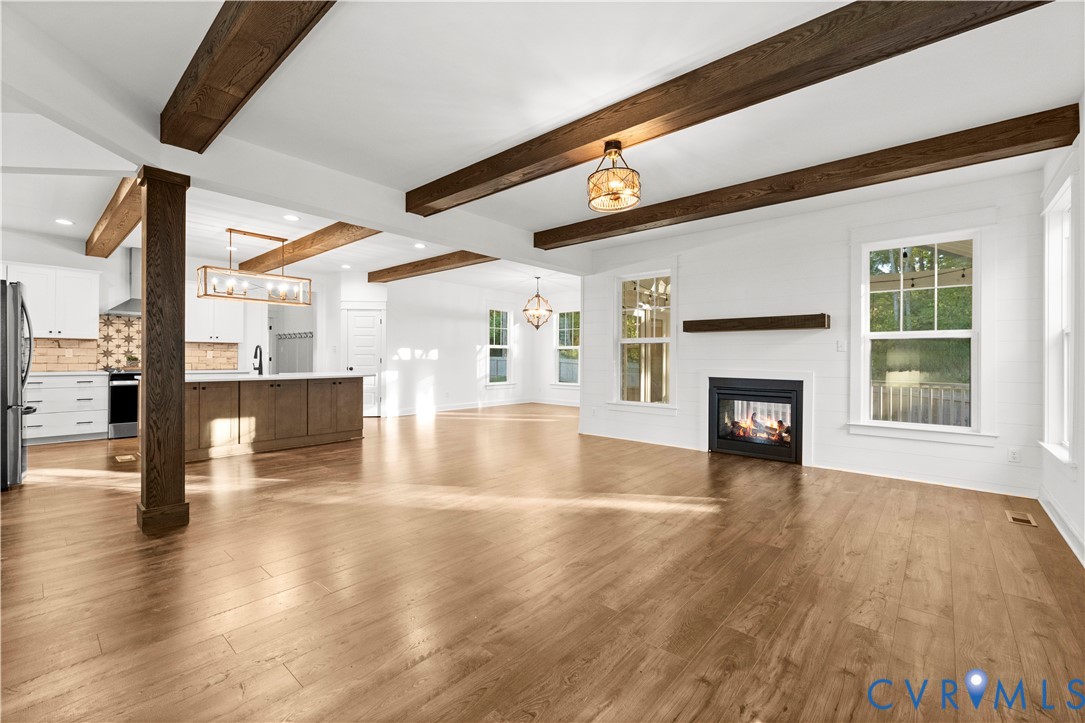 Open-concept space featuring exposed dark wood ceiling beams, wood-finish flooring, a fireplace with a wood mantel, and kitchen cabinetry with a central island