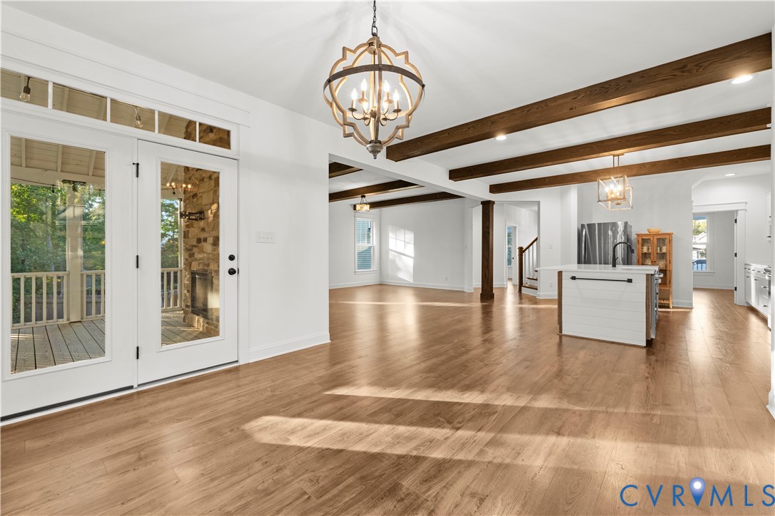 Open-concept interior featuring wood-finish flooring, exposed stained-wood ceiling beams, a decorative iron chandelier, a center kitchen island, and glass-paneled double doors with transom windows