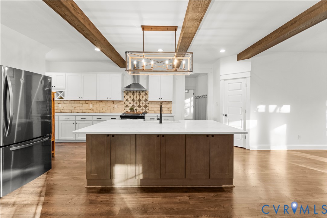 Kitchen featuring white shaker-style cabinetry, center island with stone-surface countertop, exposed stained-wood ceiling beams, patterned tile backsplash, and wood-finish flooring