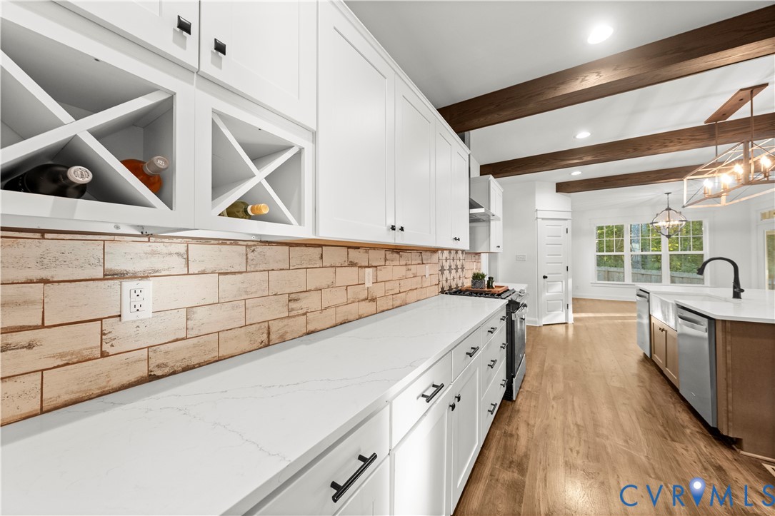Kitchen featuring white cabinetry with integrated wine storage, light-toned stone-style backsplash, wood-finish flooring, exposed ceiling beams, and a farmhouse-style sink