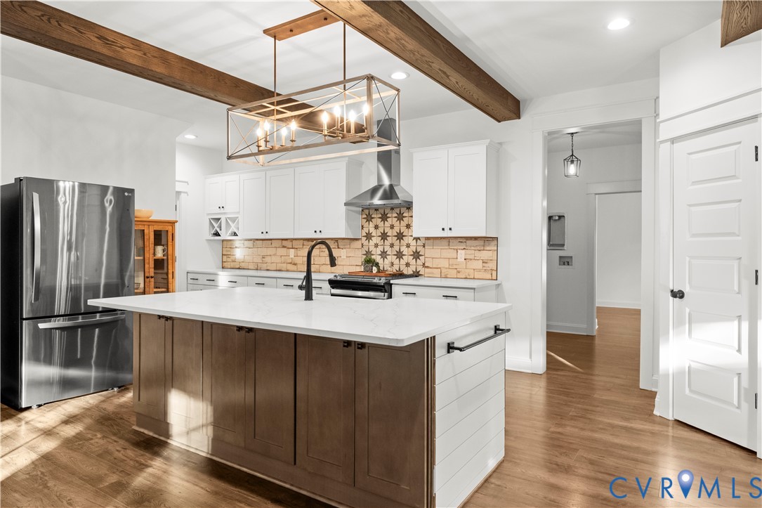 Kitchen featuring a central island with a solid surface countertop, white shaker-style cabinetry, a patterned tile backsplash, wood-finish flooring, and exposed ceiling beams