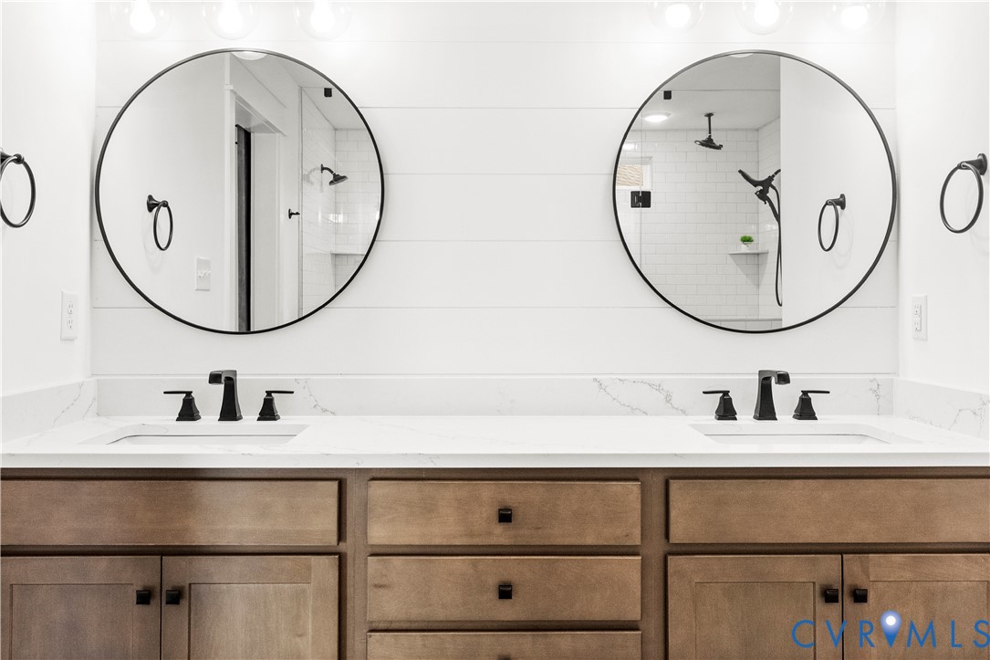 Double vanity featuring wood-finish cabinetry, white stone-composite countertops, dual circular mirrors with black frames, and matte black plumbing fixtures set against shiplap wall detailing