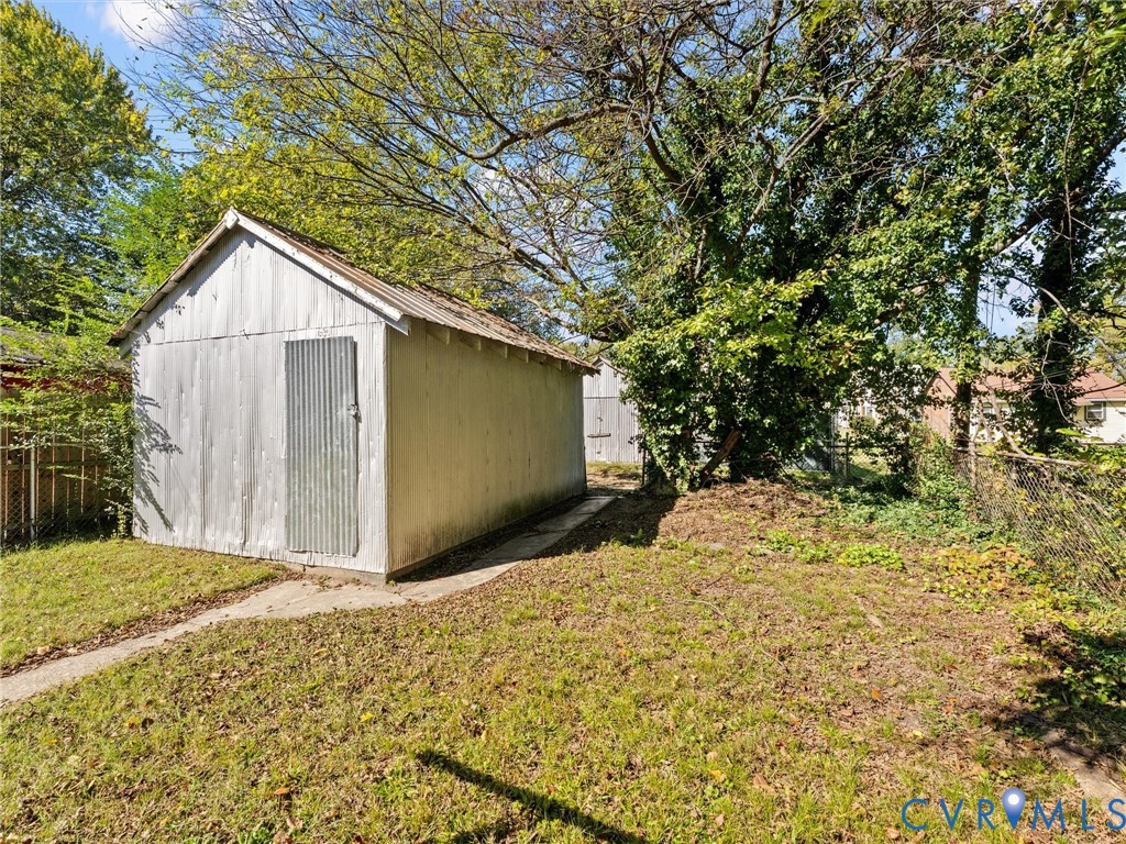 Detached metal storage structure with a corrugated finish, gable roof, and concrete pathway access on a lot with mature trees and chain-link fencing