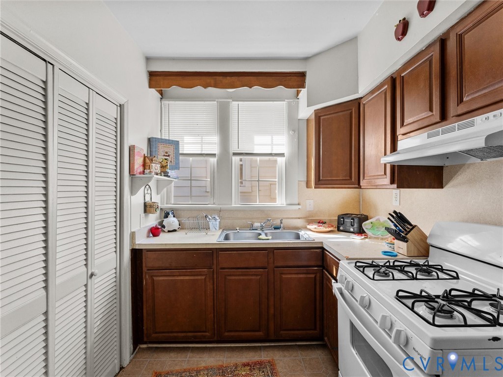 Kitchen featuring wood cabinetry, white double-door louvered pantry, dual-basin sink, and gas range with integrated vent hood