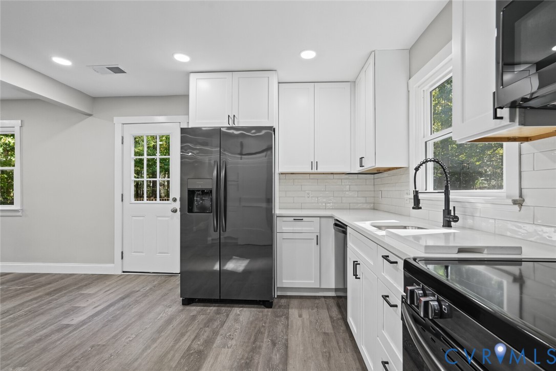 Kitchen featuring white shaker-style cabinetry, light-toned stone countertops, a subway tile backsplash, dark-finish hardware, and wood-finish flooring