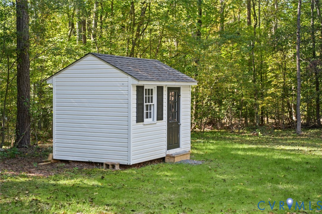 Detached outbuilding featuring white vinyl siding, a gabled roof, dark shutters, and a single-entry door, set within a grassed lot bordered by mature trees