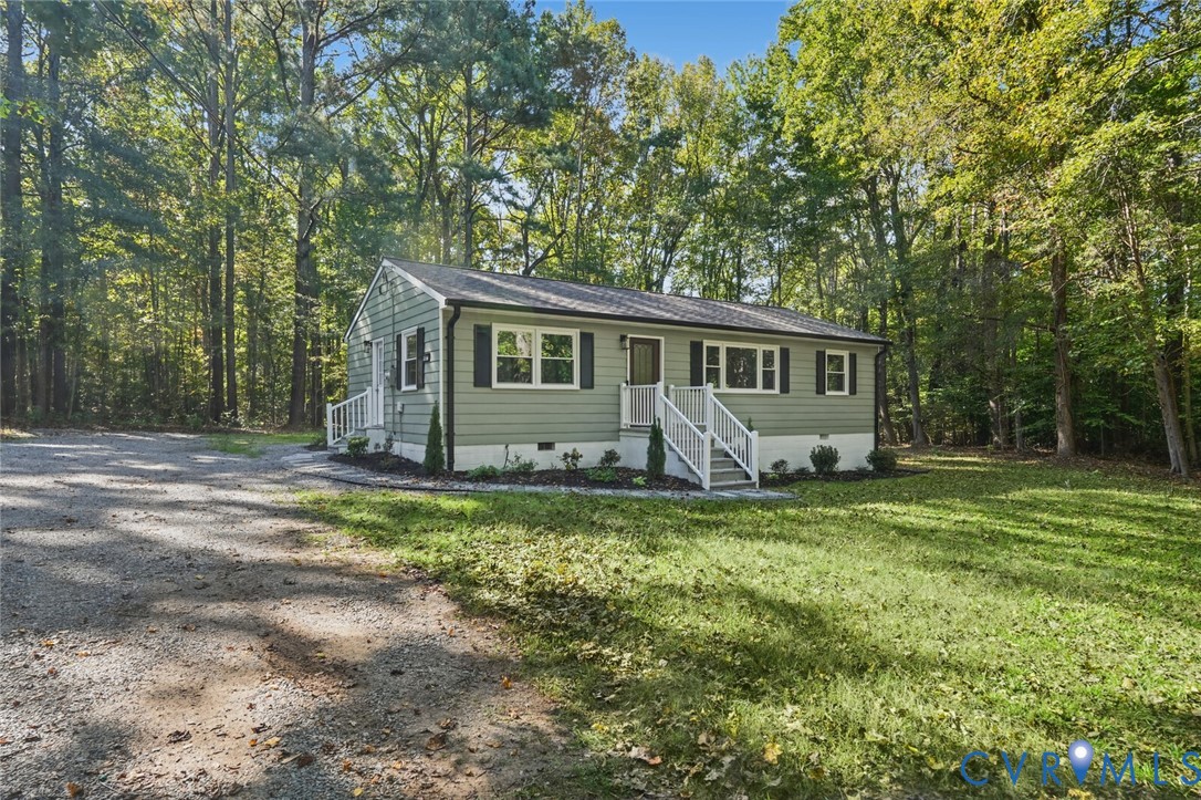 Single-story home featuring sage green siding, contrasting dark shutters, multi-pane windows, elevated white-railed entry stairs, and a gravel driveway