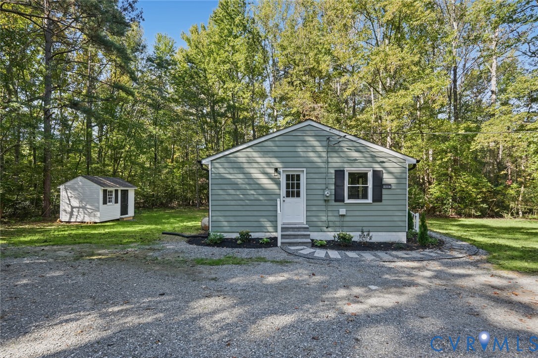 Exterior featuring light-tone horizontal siding, dark shutters, a white entry door with glass paneling, a gravel driveway, and a detached storage structure