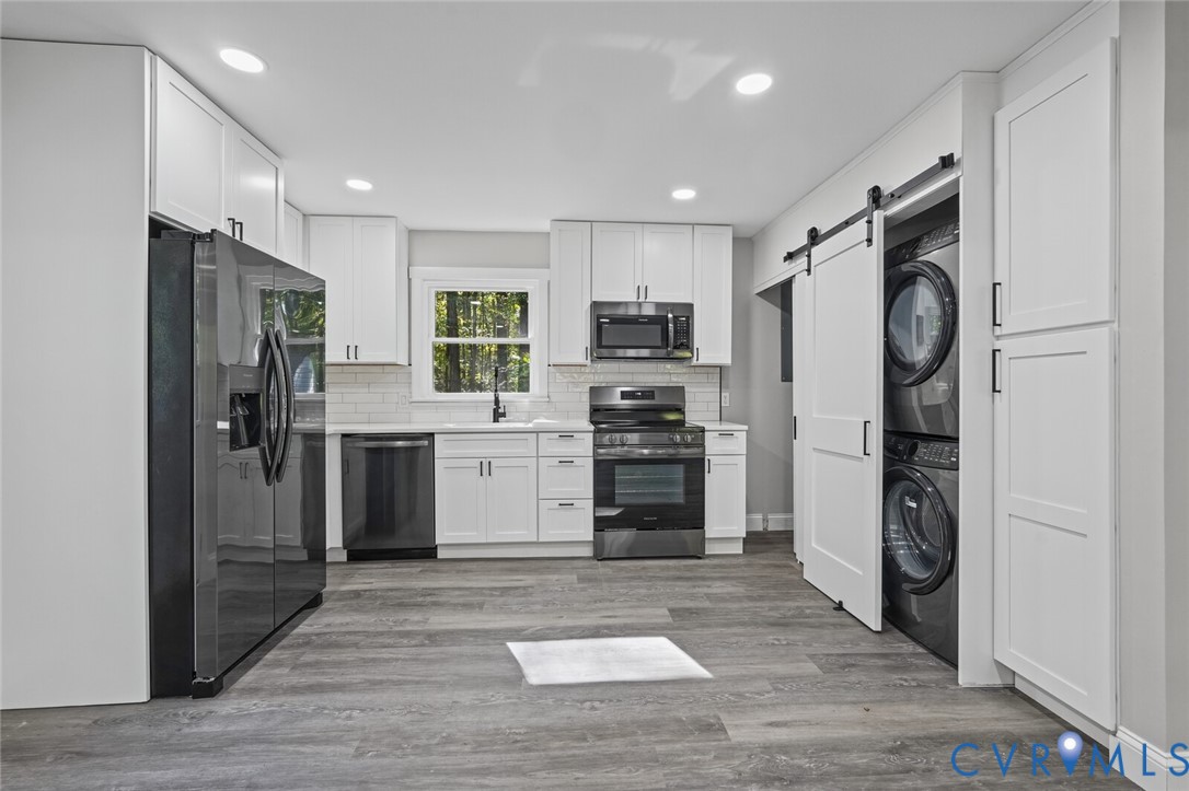 Kitchen featuring white shaker-style cabinetry, wood-finish flooring, a white subway tile backsplash, dark stainless steel appliances, and a sliding barn door concealing the laundry area
