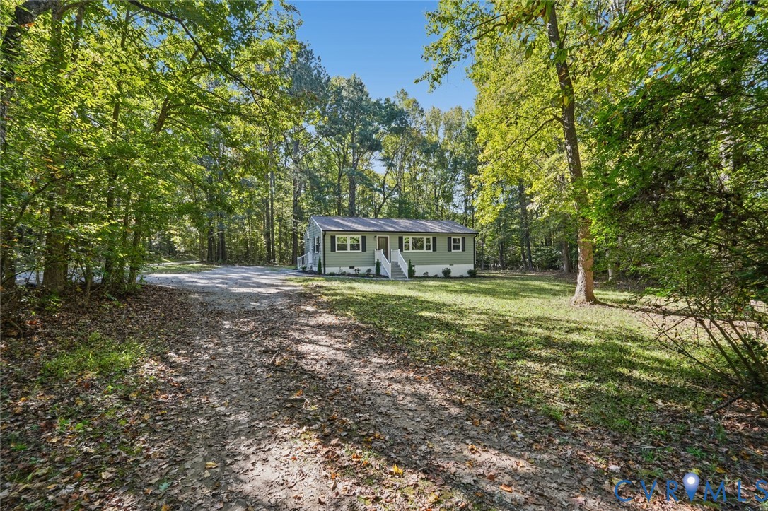 Single-story home featuring a light green exterior, white-railed front steps, shingle roof, dark shutters, and a gravel driveway situated on a wooded lot