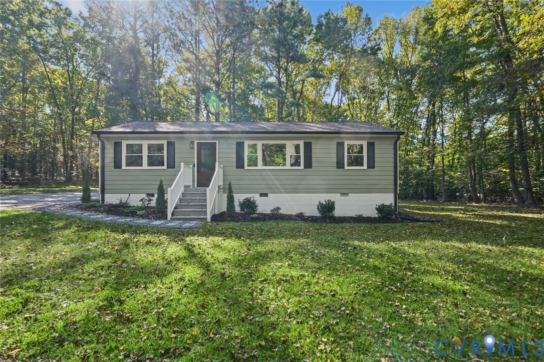 Single-story exterior featuring light-toned horizontal siding, decorative window shutters, a stone paver walkway, white railings, and mature wooded surroundings