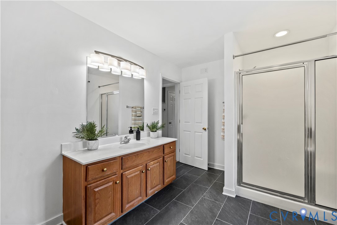 Bathroom featuring a wood-finish vanity with a stone countertop, an integrated sink, a multi-bulb light fixture above a large mirror, and a shower with sliding glass doors over dark tile flooring