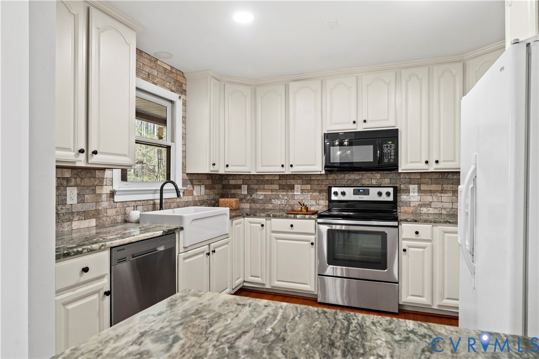 Kitchen featuring cream-colored cabinetry, stone-style tile backsplash, farmhouse-style sink with a gooseneck faucet, stone countertops, and stainless steel appliances
