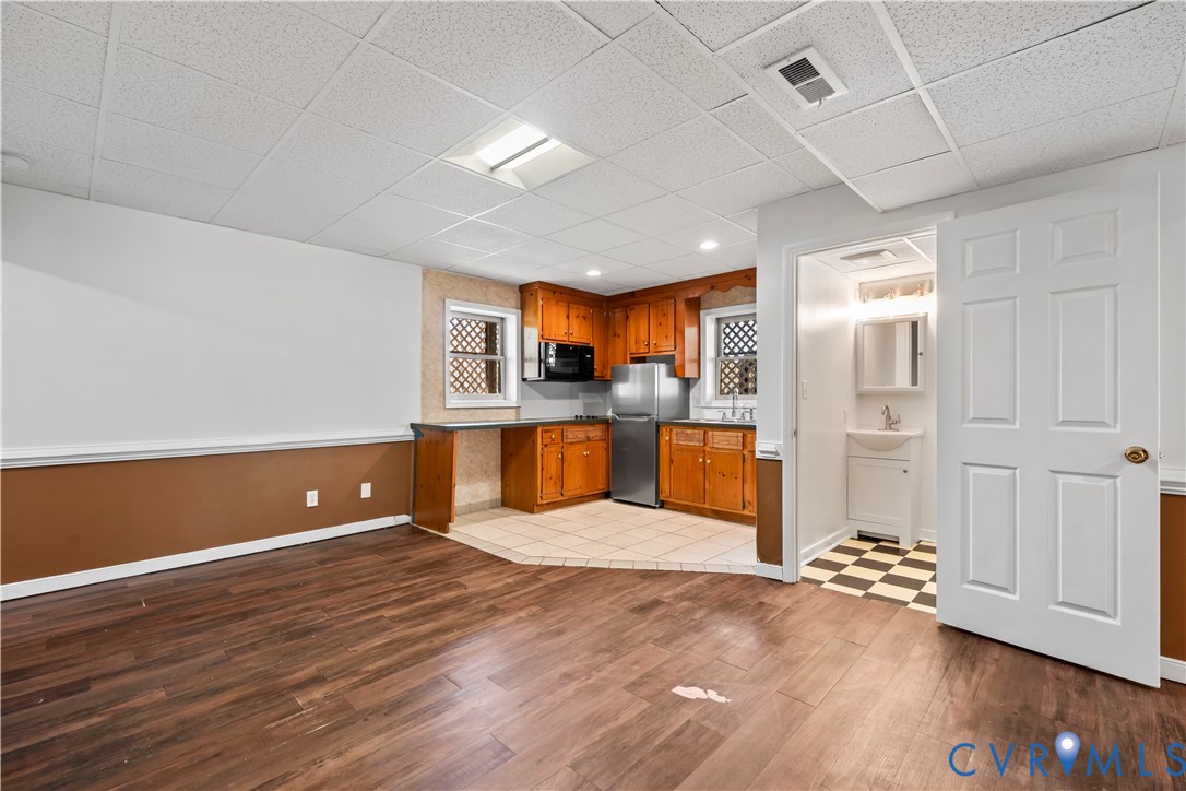Wood-finish flooring, drop ceiling with recessed lighting, kitchen featuring wood cabinetry and stainless steel appliances, ceramic tile kitchen flooring, and a private restroom with a pedestal sink