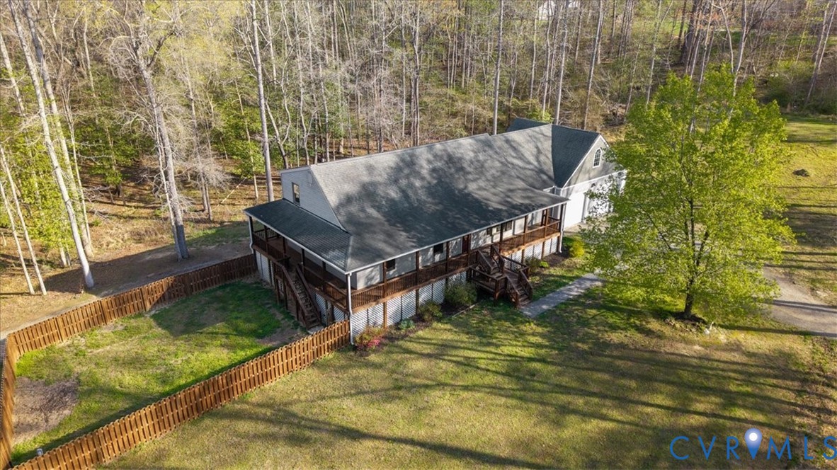 Multi-level residence featuring a wrap-around wooden deck, shingle roof, attached garage, and a fenced-in yard, bordered by mature woodland