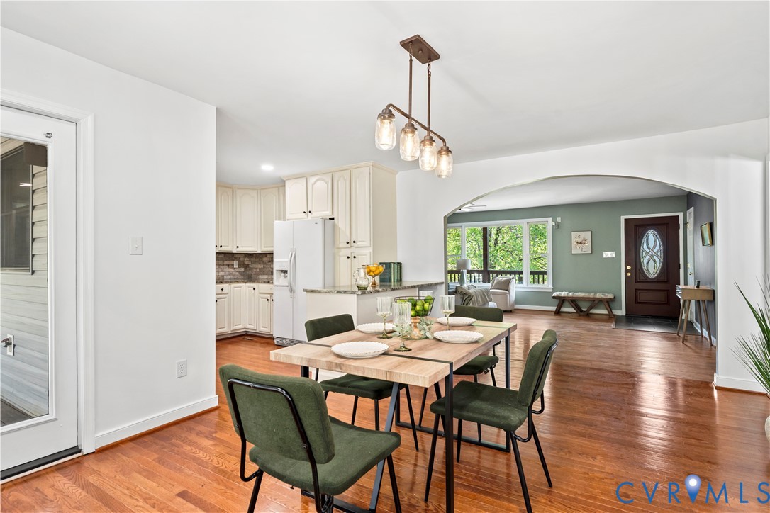 Dining area featuring wood-finish flooring, a linear four-light pendant fixture, white cabinetry, a stone-surround backsplash, and a prominent architectural archway Dining area featuring wood-finish flooring, a linear four-light pendant fixture, white cabinetry, a stone-surround backsplash, and a prominent architectural archway