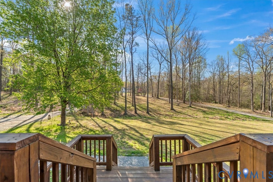Elevated wood deck featuring vertical balusters and a wide staircase, overlooking a expansive grass lot with mature trees
