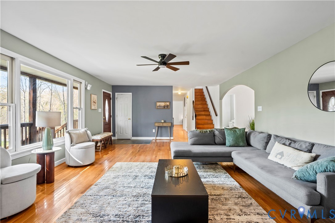 Living area featuring polished wood-finish flooring, an arched entryway, natural wood-tone staircase, ceiling fan, and large windows Living area featuring polished wood-finish flooring, an arched entryway, natural wood-tone staircase, ceiling fan, and large windows