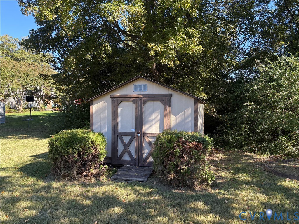 Exterior storage shed featuring double doors, a gable roof, and wood-finish trim, situated on a grassy lot with mature trees