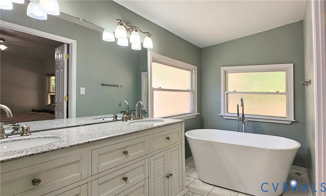 Dual-sink vanity with stone-finish countertop, white cabinetry, freestanding soaking tub, chrome fixtures, and tile flooring