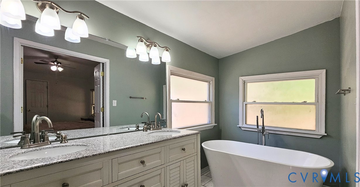 Double vanity with stone-finish countertop, undermount sinks, freestanding soaking tub, dual wall-mounted light fixtures, and large plate glass mirror