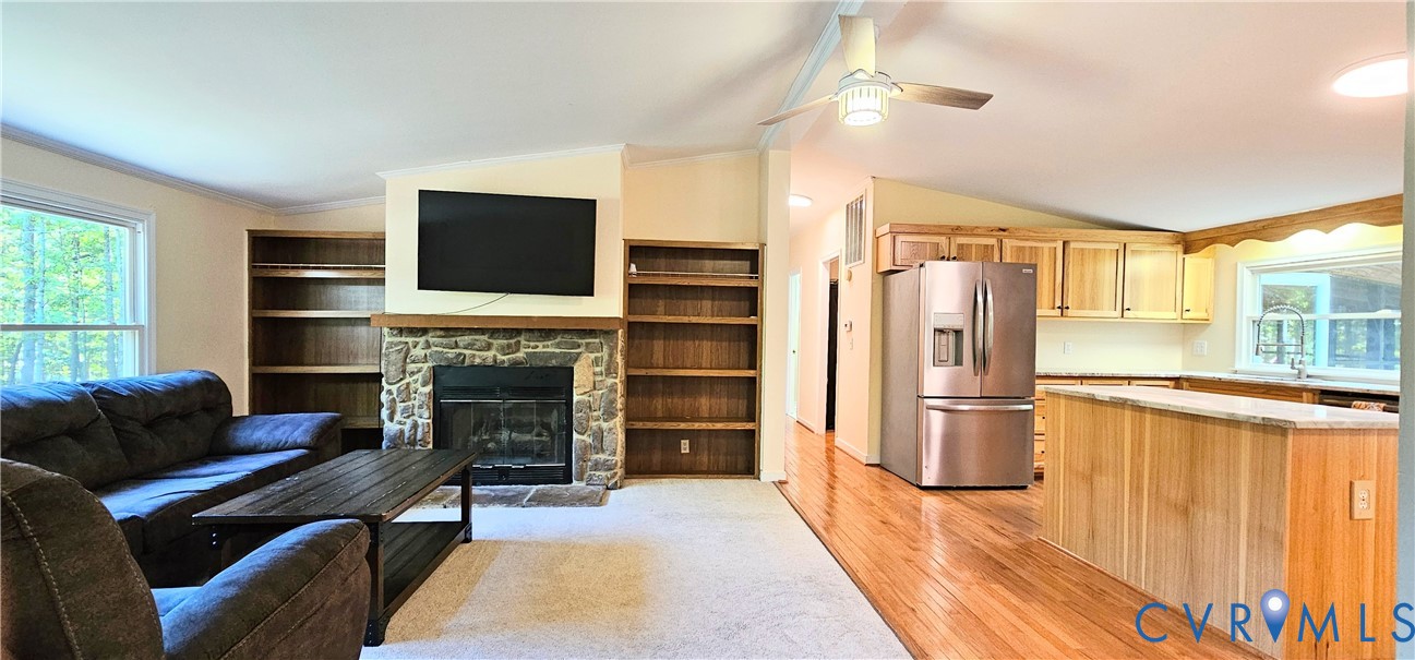 Open concept living area featuring a stone fireplace with integrated built-in shelving, hardwood flooring, wooden cabinetry, and a ceiling fan