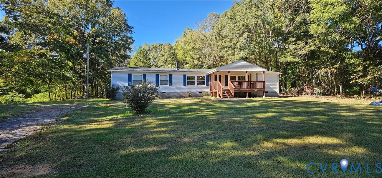 Single-story exterior featuring light-toned siding, blue window shutters, a covered wooden front deck, and expansive surrounding grounds with mature trees