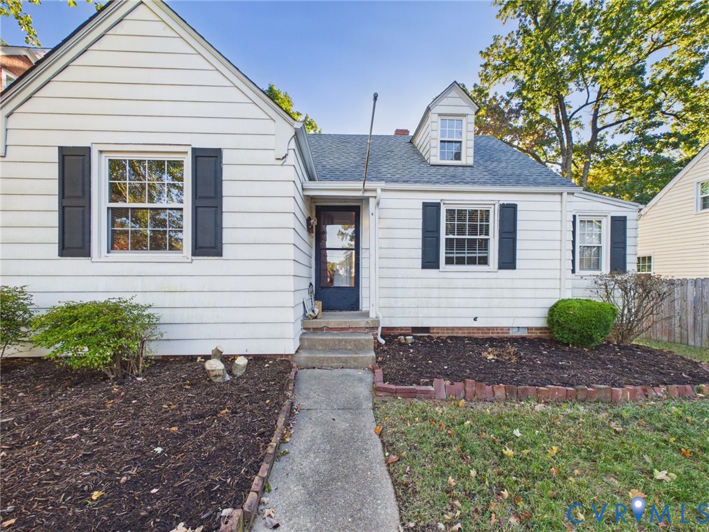 Cape Cod exterior featuring white horizontal siding, dark decorative shutters, a gabled dormer window, and a concrete walkway bordered by brick edging