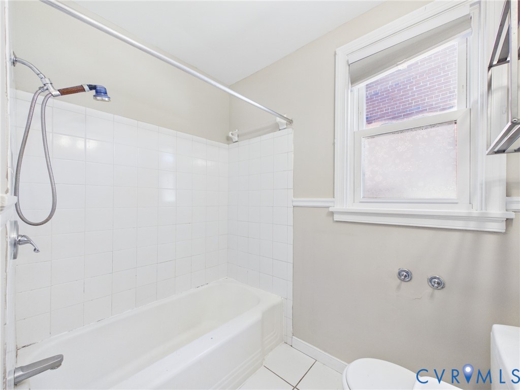 Bathroom featuring a white tub-shower combination with ceramic wall tile, a handheld shower fixture, a single-hung window with decorative trim, and tiled flooring