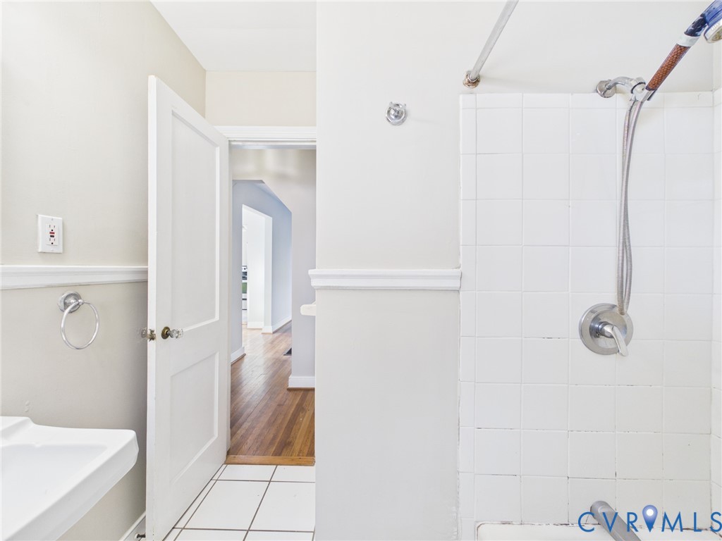 Wall-mounted hand shower fixture, white ceramic wall tiling, pedestal sink, chair rail molding, and wood-finish flooring