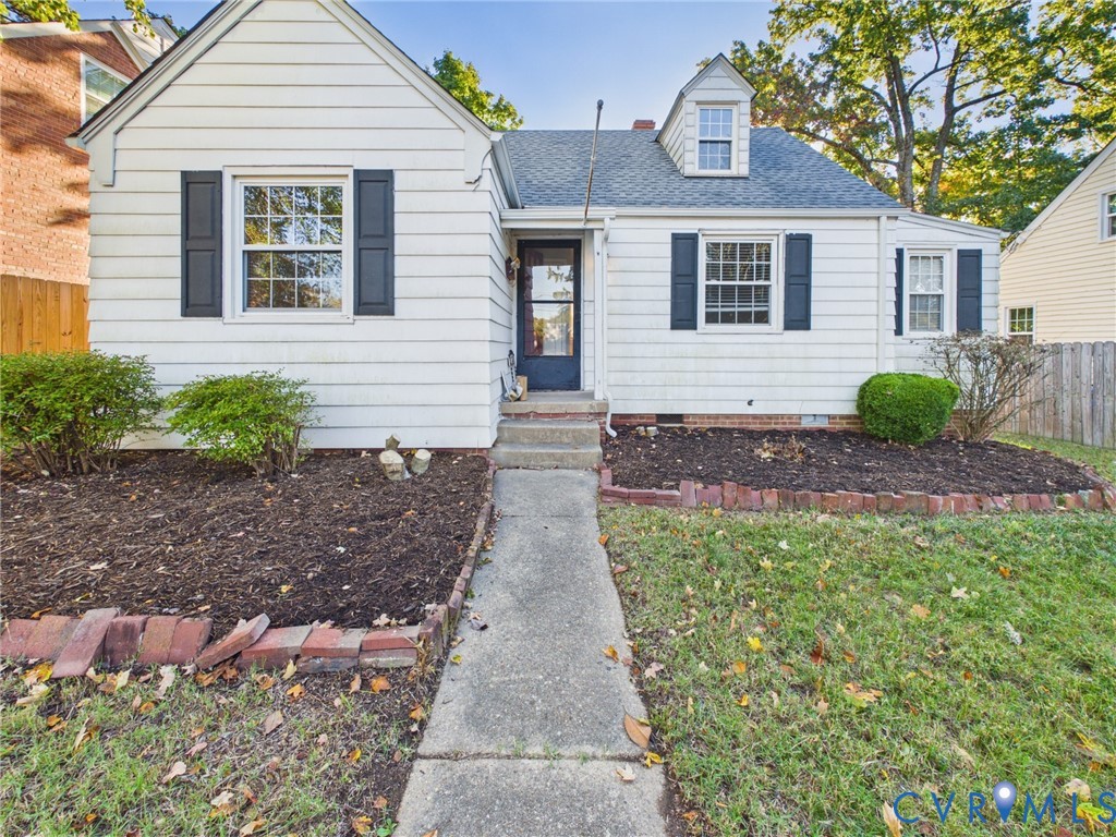 Cape Cod exterior featuring white horizontal siding, dark window shutters, a gabled roofline with a dormer, and brick-edged garden beds Cape Cod exterior featuring white horizontal siding, dark window shutters, a gabled roofline with a dormer, and brick-edged garden beds