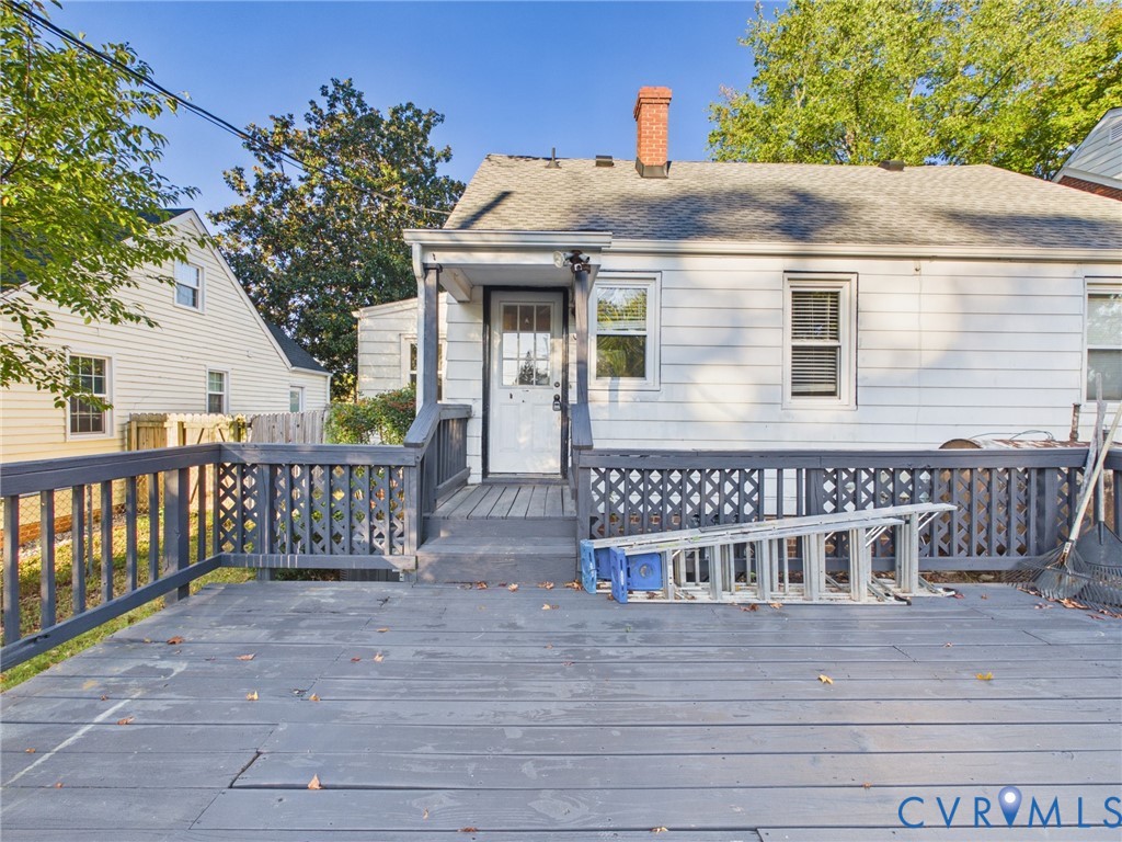 Exterior rear elevation featuring white siding, a grey composite deck with lattice railings, a masonry chimney, and multi-pane glass entry door