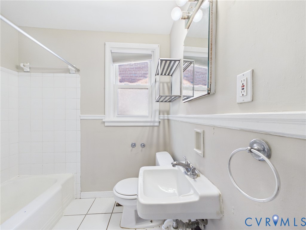 Bathroom featuring a white porcelain wall-mounted sink with a metallic faucet, white tile floor, tub with a tiled surround, and wall-mounted vanity mirror with integrated lighting