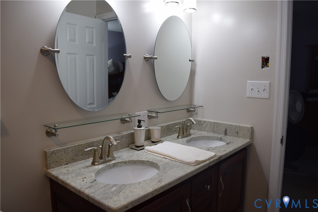 Dual-sink vanity featuring a granite countertop, oval mirrors, glass shelving, and dual-sink custom-height vanity.